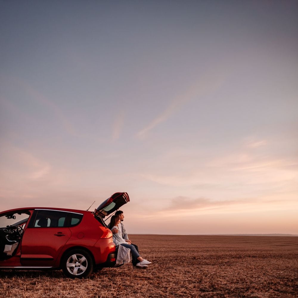 A couple sitting in the open boot of a red hatchback in an open field, watching a sunset