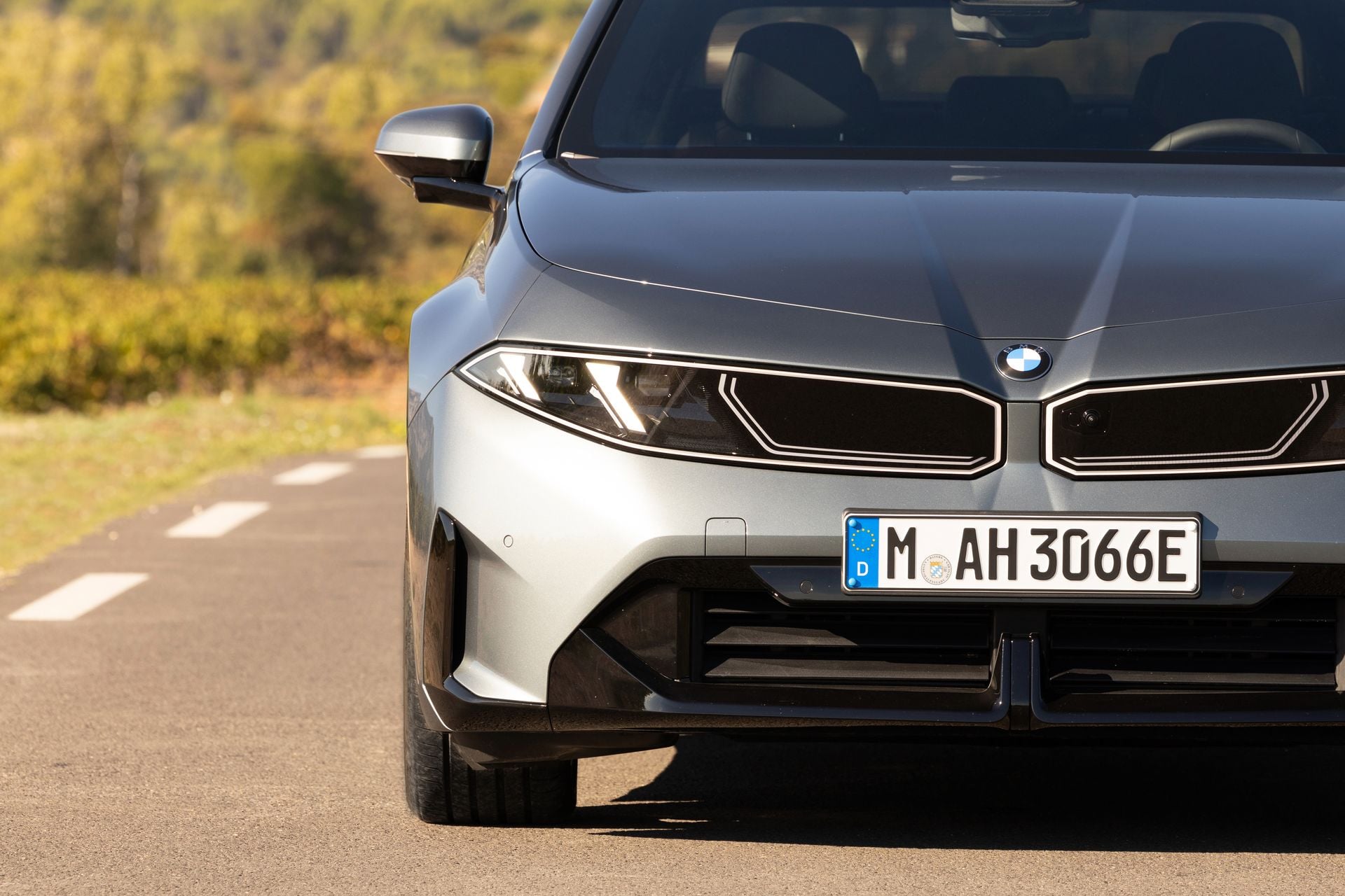 Front of a grey 2027 BMW i3 sedan showing its slim headlights, illuminated kidney grille and badge, parked on a country road