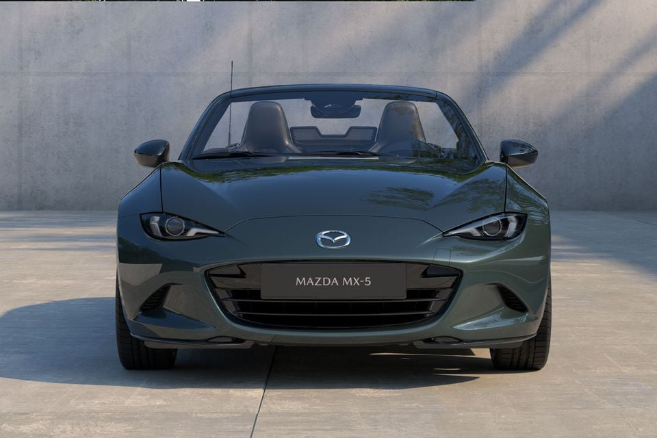 Front view of a dark green Mazda MX-5 roadster with the soft top down, photographed against a concrete wall