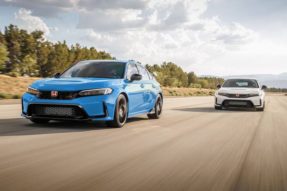 A blue Honda Civic Type R hatchback leads a white Civic Type R on a test track road.