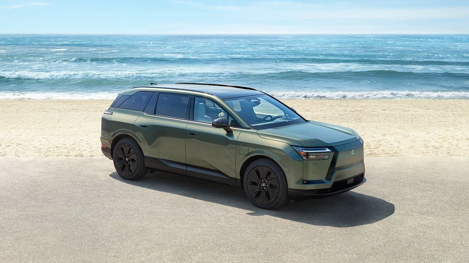 A green Lexus electric SUV seen from the front-side angle, parked on a beach with the ocean in the background