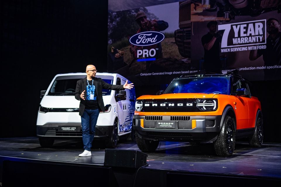 A presenter on stage at a Ford event gesturing toward two prototype vehicles — an orange Ford Bronco Basecamp and a white Ford Transit City — with a Ford Pro banner and 7-year warranty signage in the background.