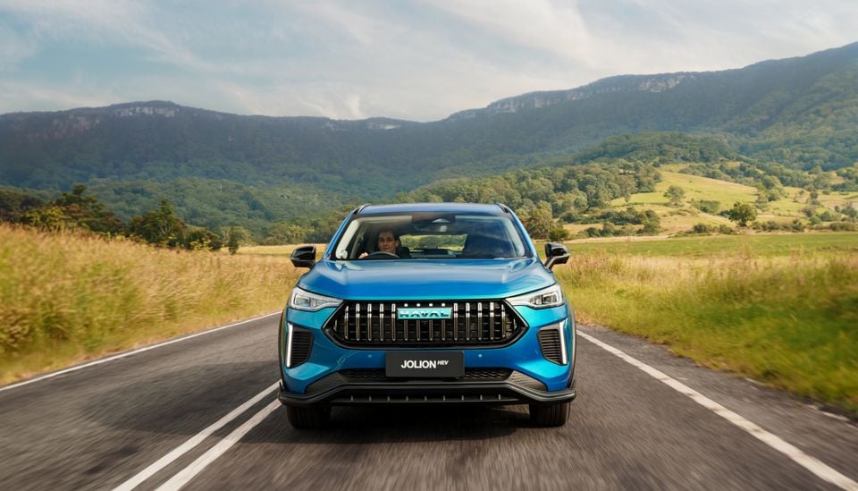 A blue Haval Jolion HEV SUV driving on a country road, seen from the front, with mountains and green fields in the background