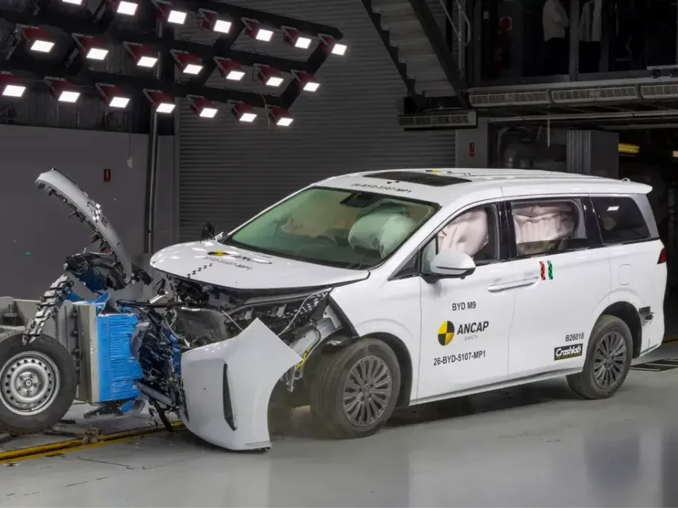A white BYD M9 people mover with a crushed front end and deployed airbags after an ANCAP frontal crash test, beside the barrier trolley