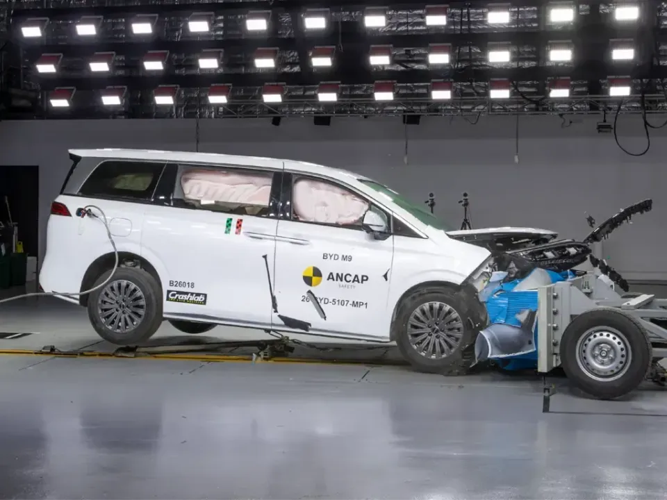 White BYD M9 people mover after an ANCAP frontal offset crash test, with deployed curtain airbags and a crushed front end against a barrier trolley