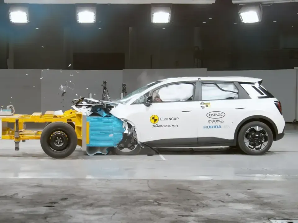 A white GAC Aion UT hatchback undergoing a Euro NCAP frontal offset crash test, striking a moving barrier with airbags deployed