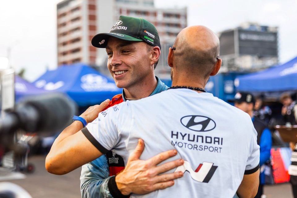 A driver wearing a Hyundai cap embraces a Hyundai Motorsport team member at a motorsport event