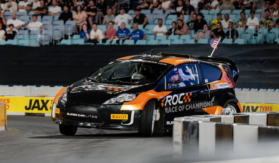 A Super Car Lites race car with ROC Race of Champions livery driven by T. Pastrana navigates a tight corner at an indoor motorsport event in front of a stadium crowd.