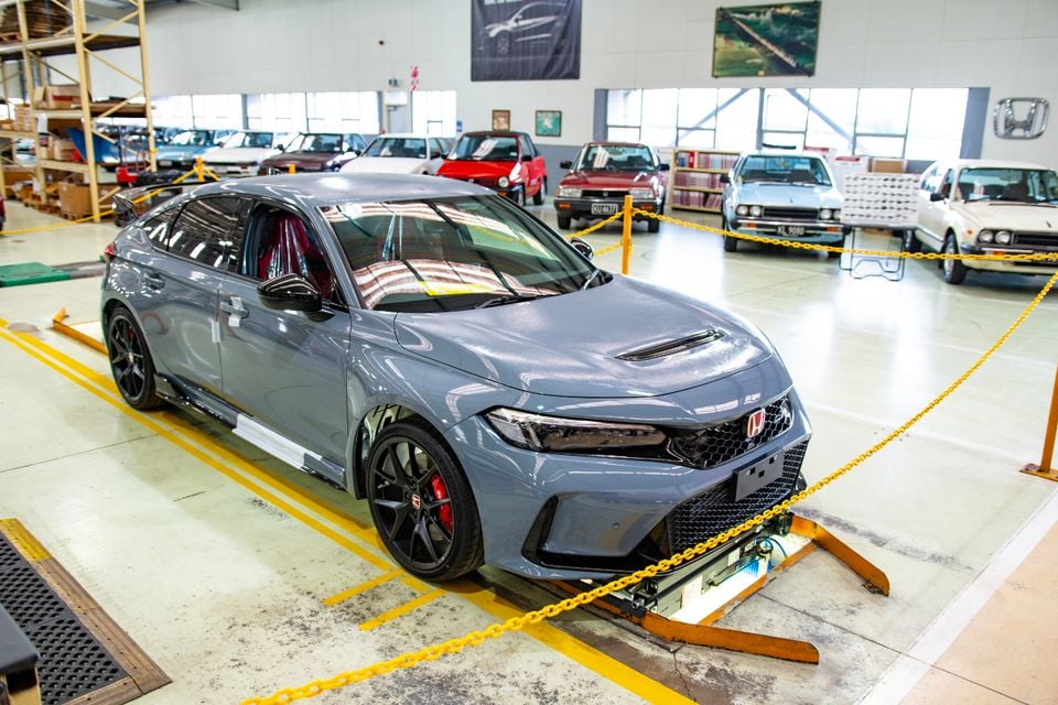 A grey Honda Civic Type R hatchback on display inside a Honda heritage facility, surrounded by classic Honda vehicles, seen from the front-side angle.