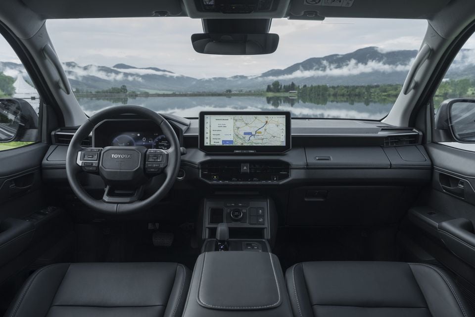 Toyota Hilux BEV interior showing the dashboard, large central touchscreen displaying navigation, steering wheel, and front seats with a scenic mountain and lake view through the windscreen