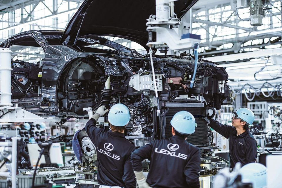 Three Lexus workers in hard hats and branded uniforms assembling a car on a production line at a Lexus manufacturing facility.