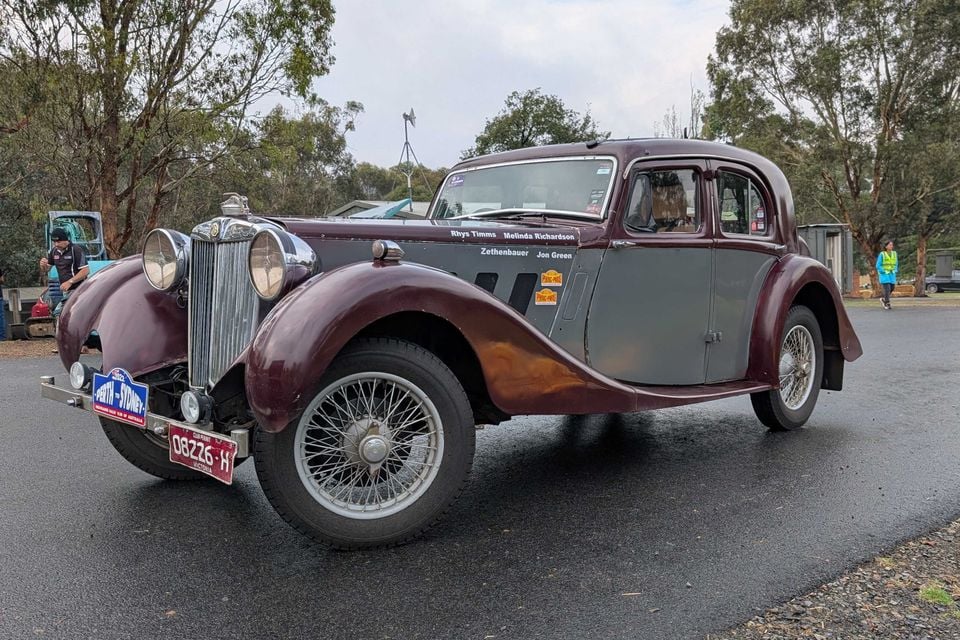 A maroon and grey 1936 MG SA saloon with wire wheels and Perth to Sydney rally plates parked on a wet road, seen from the front quarter