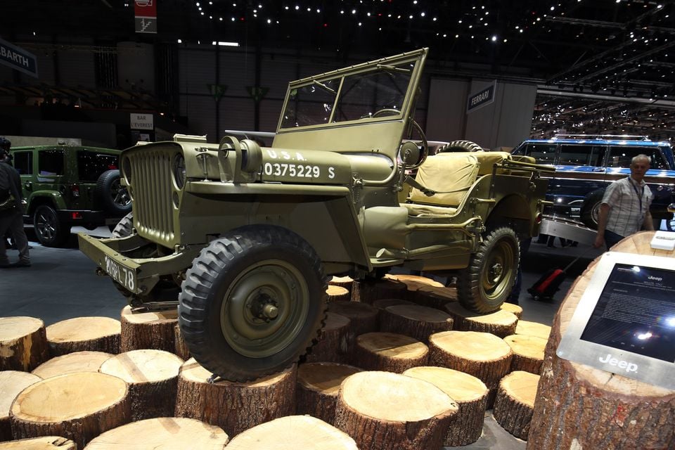 An olive green 1941 Willys Jeep military vehicle seen from the front, displayed on tree stumps at a motor show