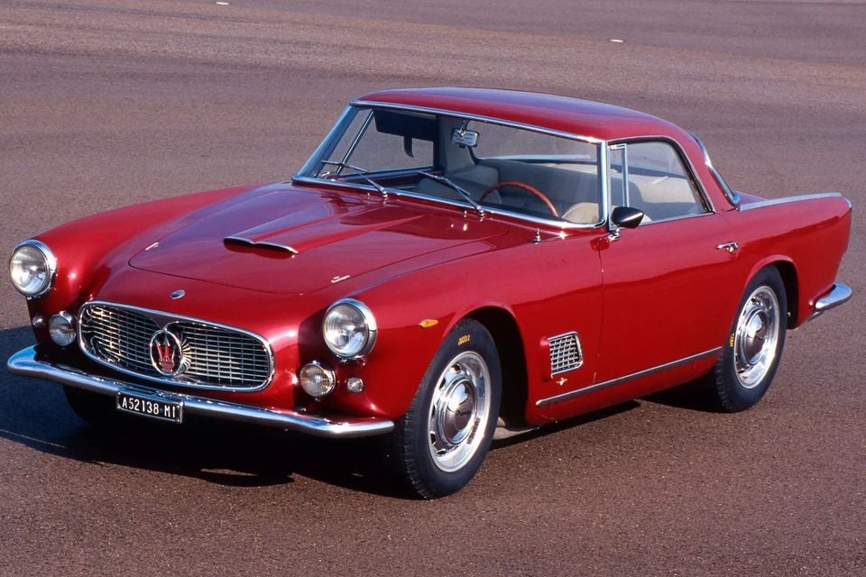 A red 1957 Maserati 3500 GT coupe parked on tarmac, seen from the front left at an elevated angle