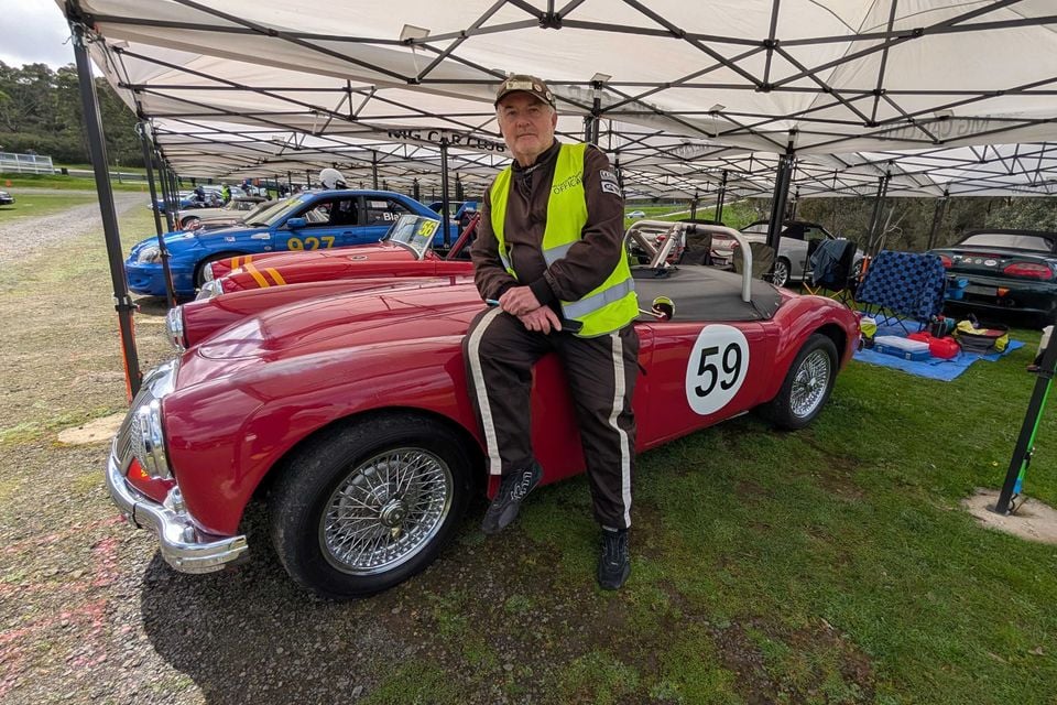 A man in a racing suit and official's vest sits on a red 1959 MGA roadster wearing race number 59, parked under an MG Car Club marquee
