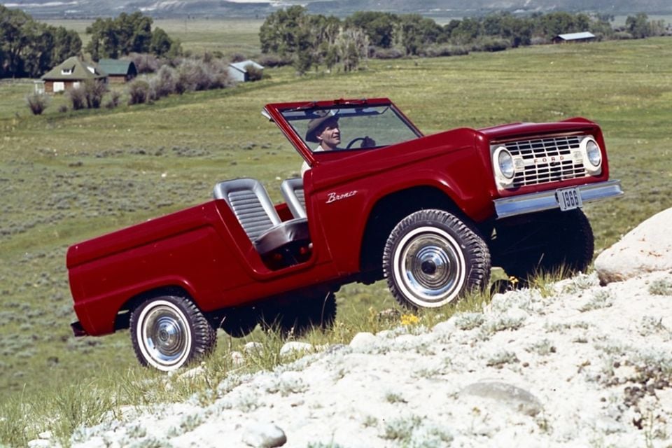 A red 1966 Ford Bronco roadster with no roof being driven up a rocky hillside in a rural landscape