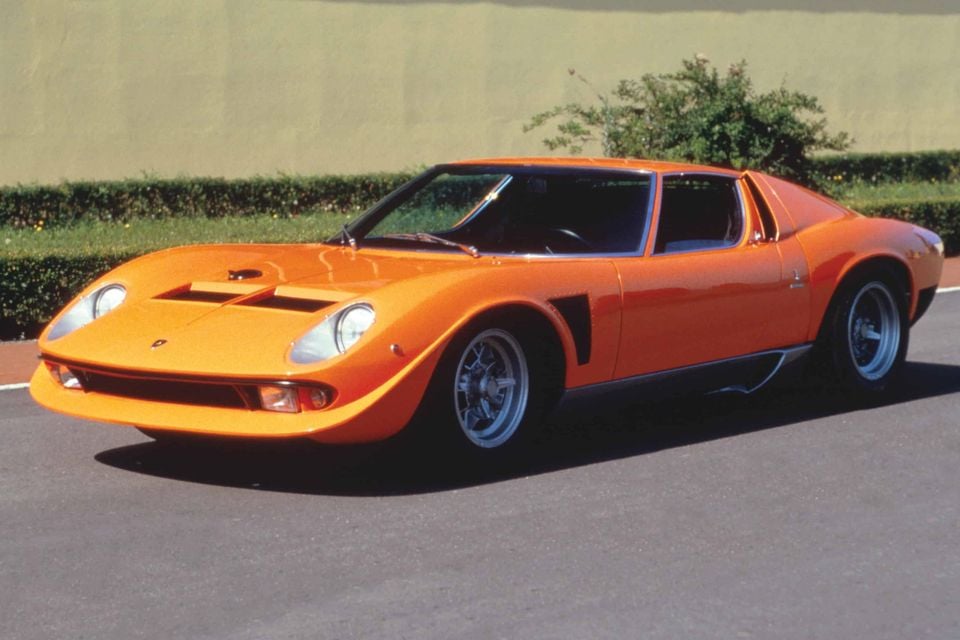 An orange 1971 Lamborghini Miura Jota supercar seen from the front-side on a road, with a hedge and wall in the background.