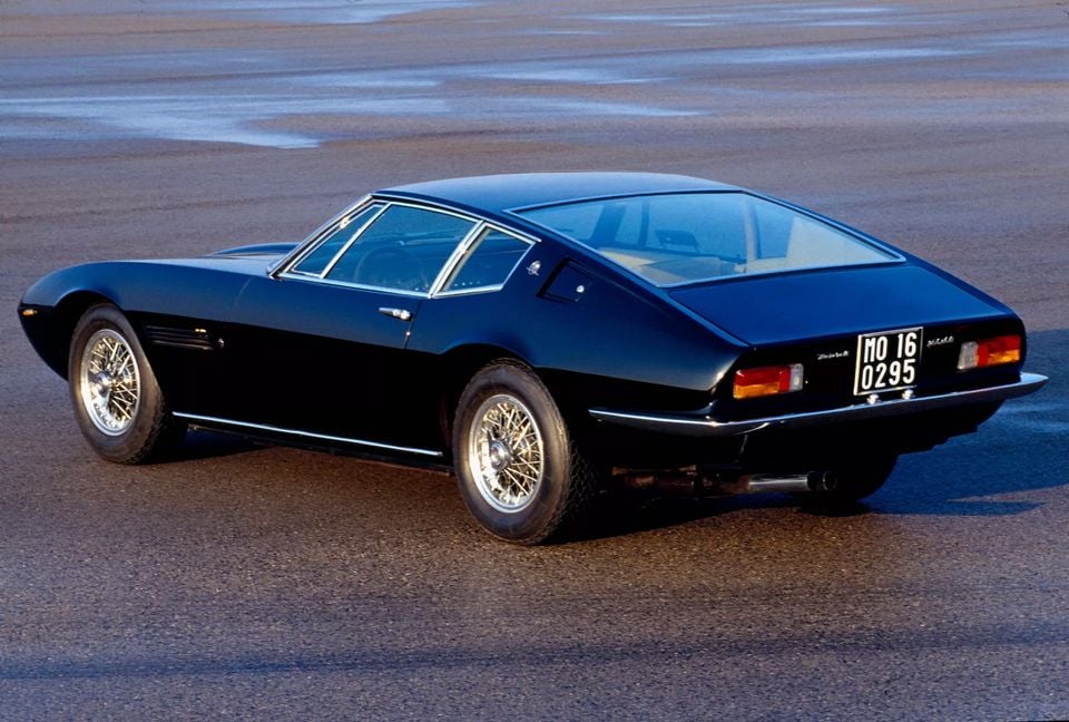 A dark blue 1971 Maserati Ghibli coupe with chrome wire wheels parked on tarmac, seen from the rear and side