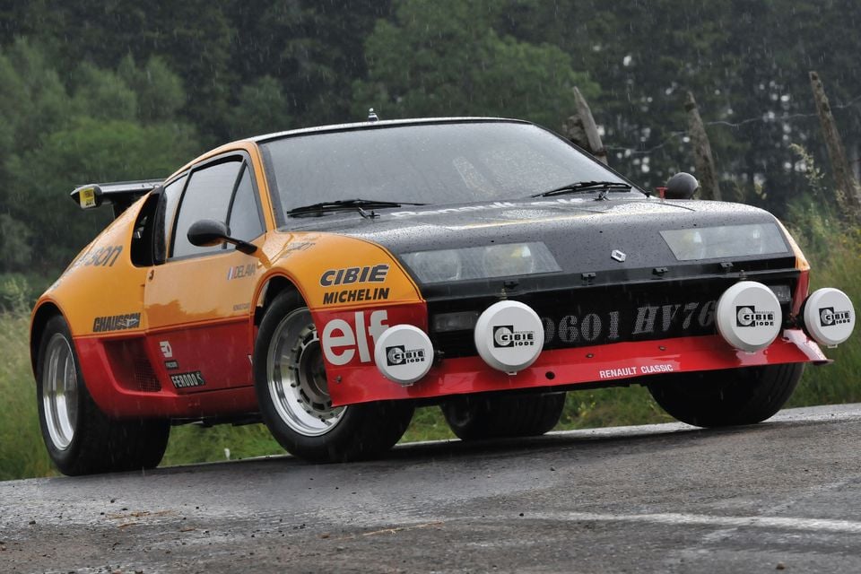 Um carro de rally Alpine A310 Grupo B 1977 com pintura Elf laranja, vermelha e preta com luzes de direção Cibie, estacionado em uma estrada molhada na chuva