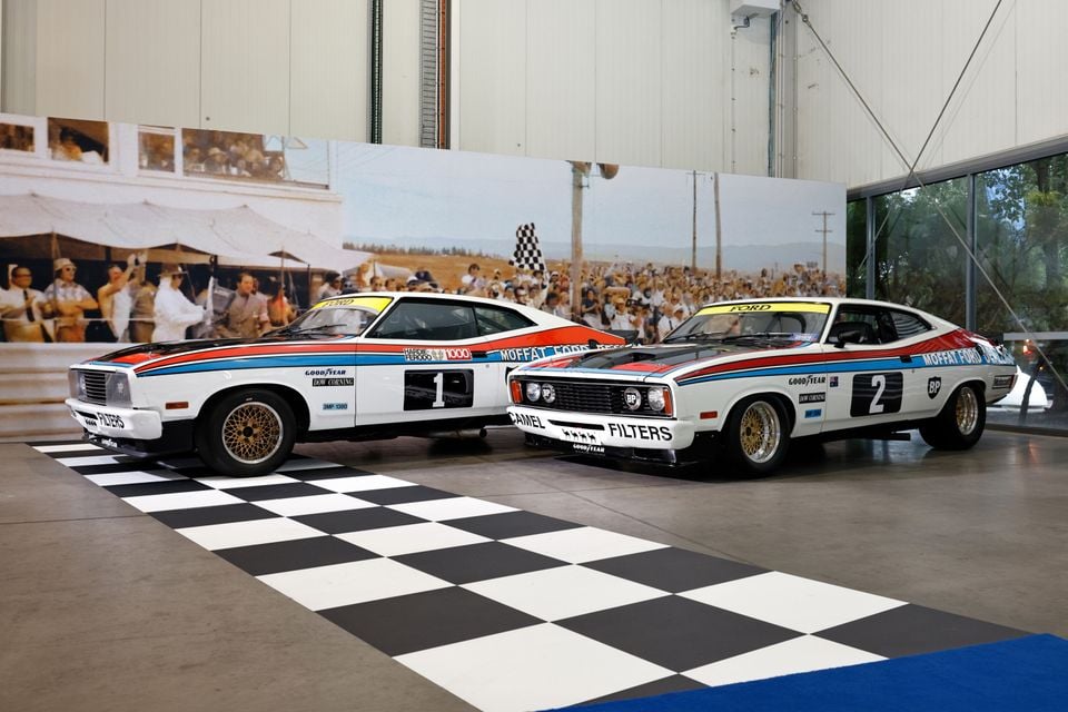 Two classic Ford Falcon XC Bathurst touring cars, numbered 1 and 2 with Moffat Ford Dealers livery, displayed on a chequered floor in a showroom.