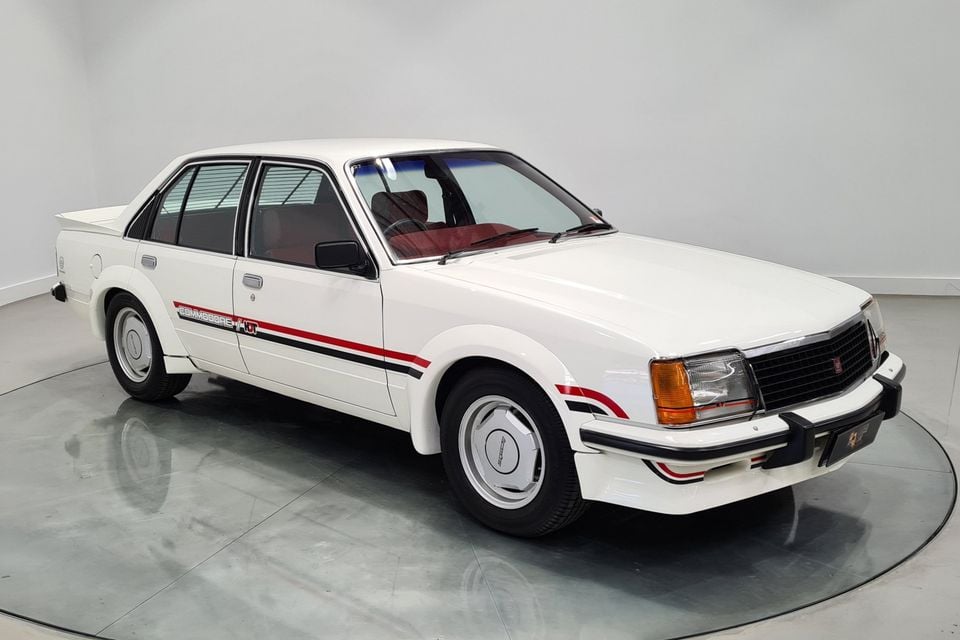 A white 1980 Holden Dealer Team Commodore VC sedan with red and black stripes, parked on a turntable in a showroom, seen from the front three-quarter