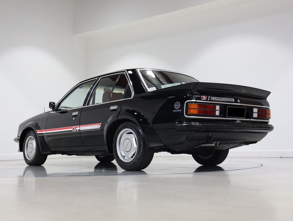 A black 1980 Holden Dealer Team VC Commodore sedan with red and white HDT side stripes, seen from the rear quarter in a white studio