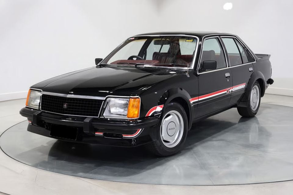 A black 1980 Holden Commodore VC HDT sedan with red and white side stripes, seen from the front three-quarter on a showroom turntable