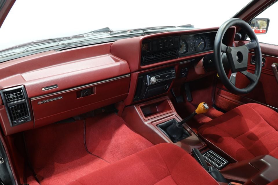 Red velour interior of a 1980 Holden Dealer Team VC Commodore, showing the dashboard, steering wheel, manual gear shifter and front seats