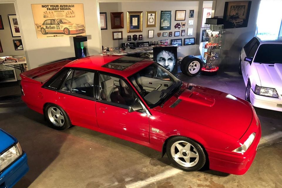 A red 1986 Holden Dealer Team VL Commodore SS Group A sedan seen from above at the front, parked in a garage filled with racing memorabilia