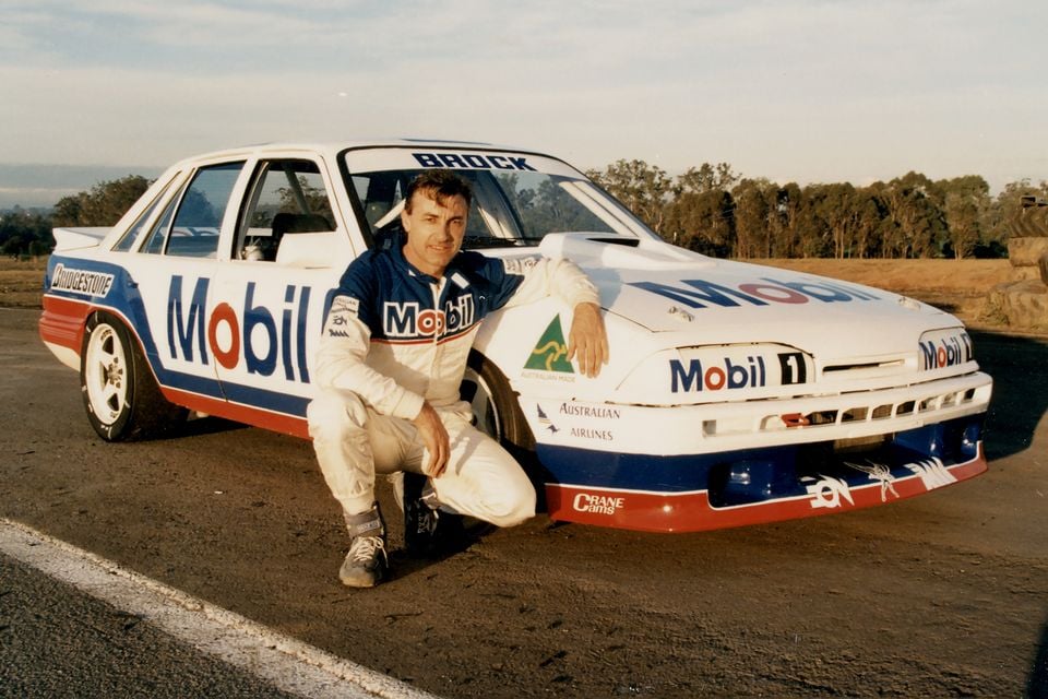 Peter Brock crouches beside his Mobil 1-liveried Holden VL Commodore SS Group A racing car on a track, wearing his race suit