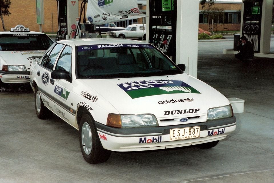 A white 1992 Ford Falcon EB sedan covered in sponsor decals, parked at a petrol station during the Eco Challenge economy run.