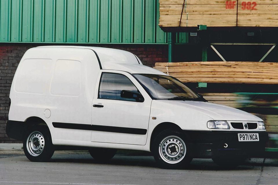 A white 1996 SEAT Inca panel van parked outside an industrial building with stacked timber in the background