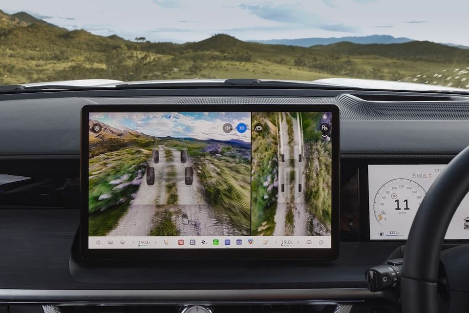 The infotainment screen of a GWM Tank 500 shows a split-screen off-road camera view of a rocky dirt trail with hills in the background.