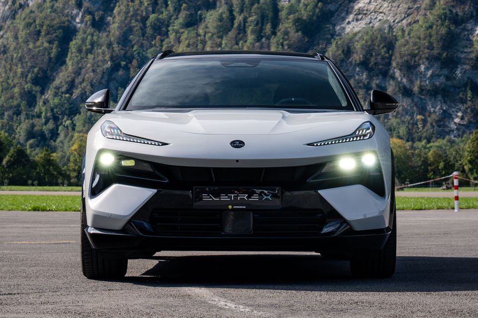 A white 2027 Lotus Eletre X SUV seen from the front with its lights on, parked on tarmac in front of a forested mountainside.