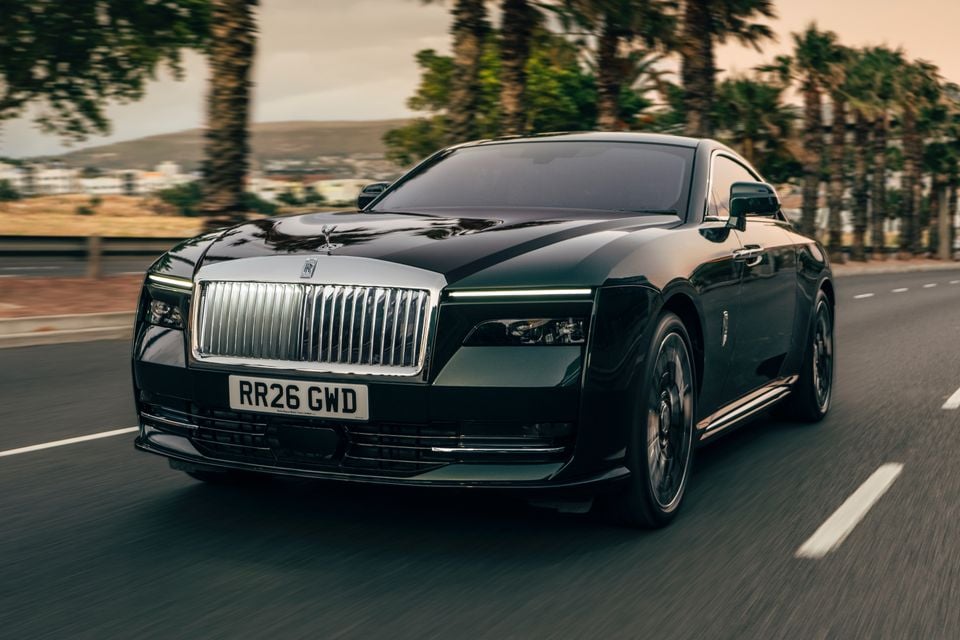 A black 2027 Rolls-Royce Spectre electric coupe driving along a palm-lined road, seen from the front