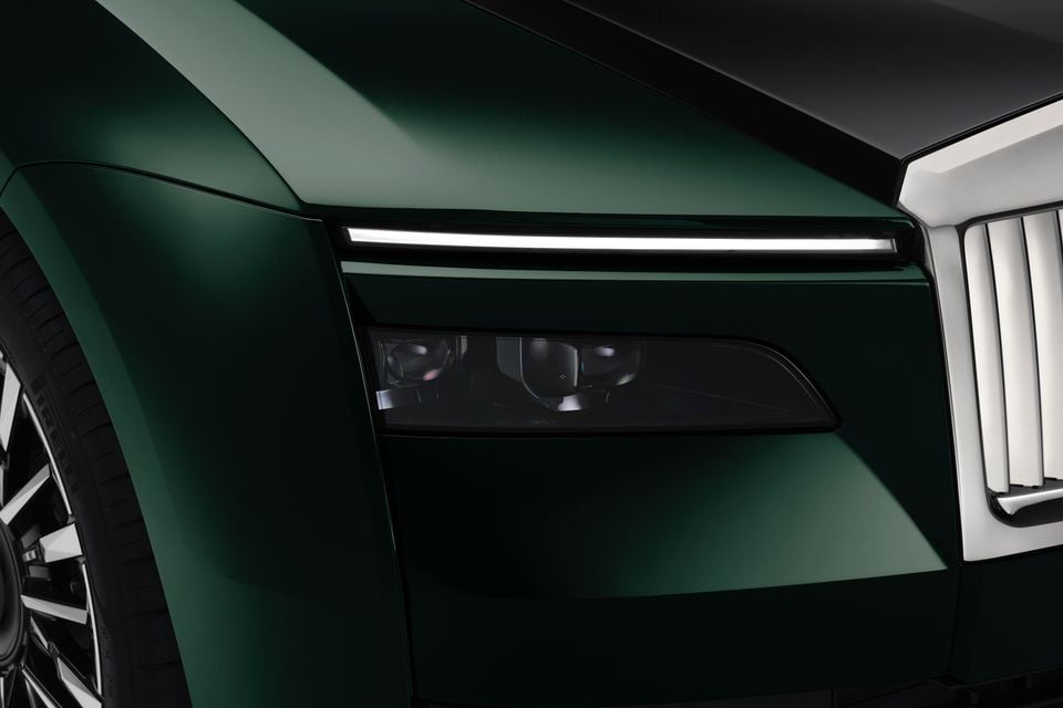 Close-up of the slim LED daytime running light and headlight on a dark green 2027 Rolls-Royce Spectre beside its illuminated grille