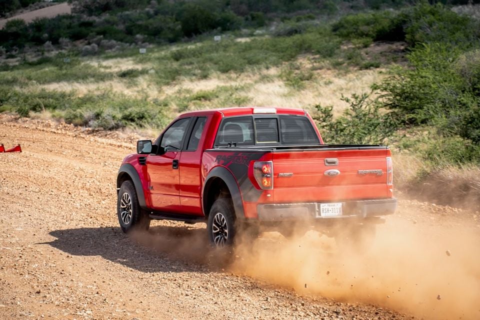 A red Ford F-150 SVT Raptor ute kicking up dust on a dirt track, seen from the rear three-quarter angle