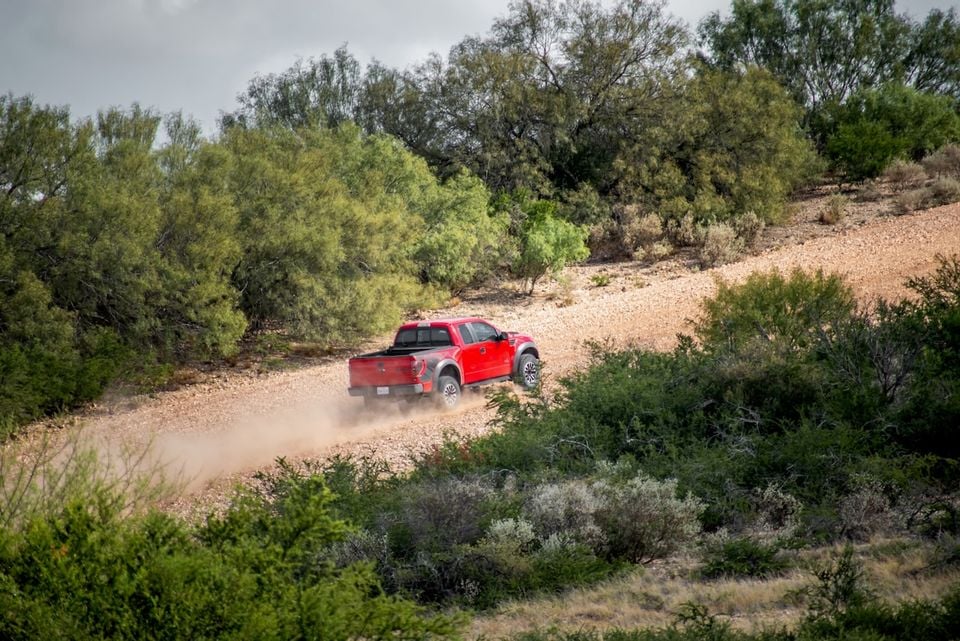 A red Ford F-150 SVT Raptor ute driving along a dusty dirt track surrounded by scrubland, seen from the rear.