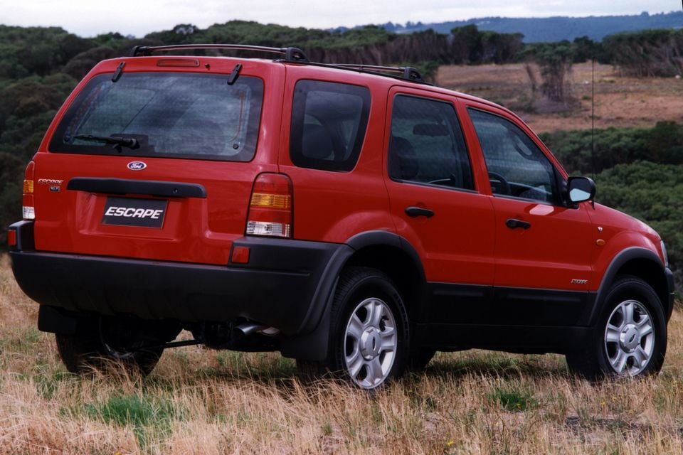 A red Ford Escape SUV seen from the rear three-quarter angle, parked on dry grassland with trees and hills in the background