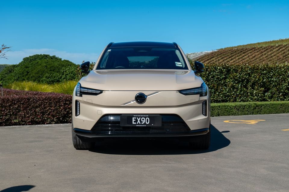 A sand-coloured Volvo EX90 large SUV seen from the front, parked in a sunny outdoor setting with hedges and vineyard rows in the background.