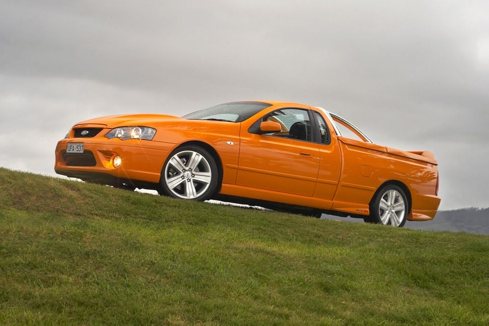 An orange 2005 Ford Falcon BF XR6 ute parked on a grassy hill, seen from the front side angle.