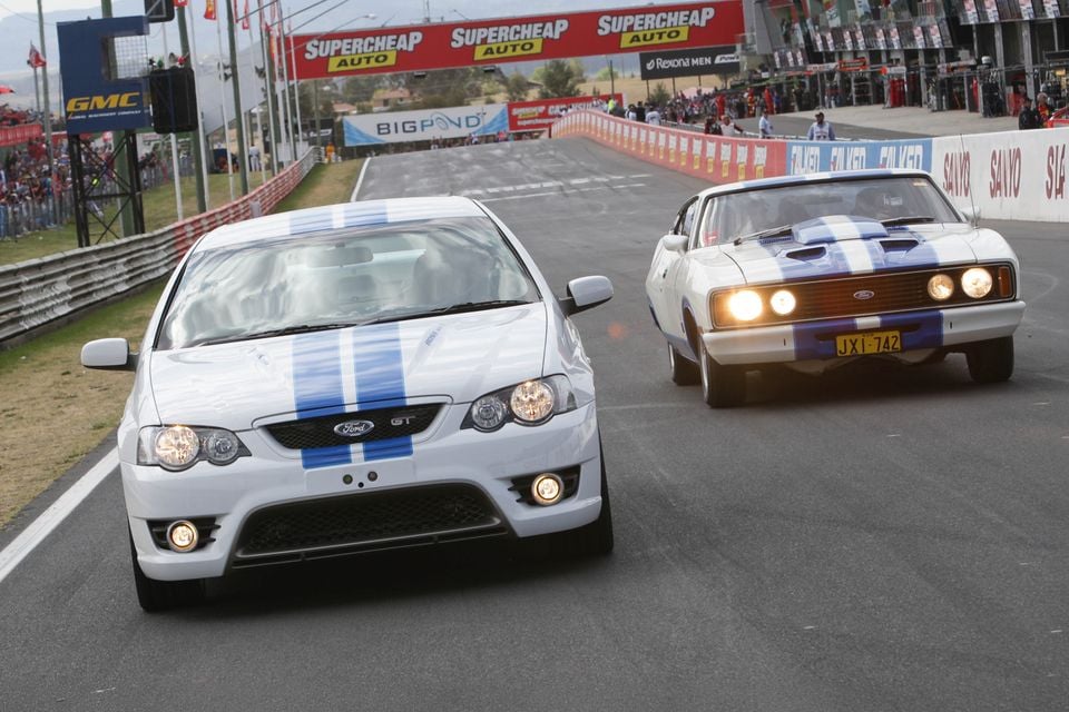 Two white Ford FPV GT performance cars with blue racing stripes, a modern model and a classic Falcon coupe, driving on Mount Panorama racetrack at Bathurst
