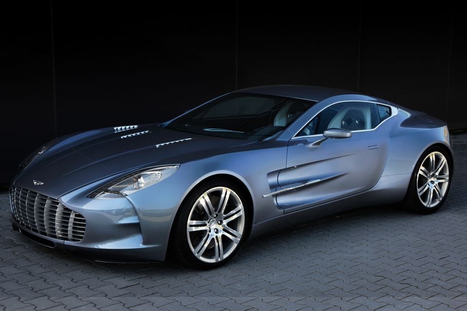 A silver-blue 2009 Aston Martin One-77 supercar coupe seen from the front-side angle, parked on a cobblestone surface against a dark background.