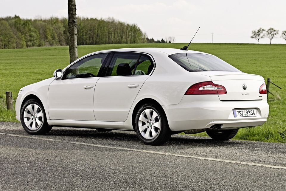 A white Skoda Superb sedan seen from the rear three-quarter angle, parked on a rural road surrounded by green fields and trees.