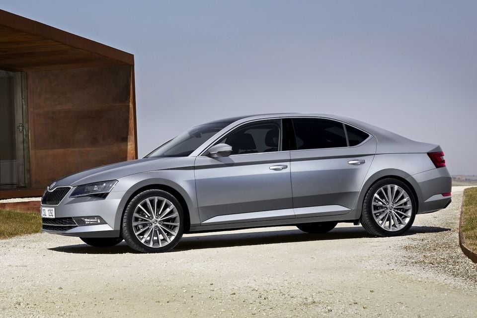 A silver Skoda Superb liftback sedan seen from the front side, parked on a gravel surface next to a modern timber-clad building.