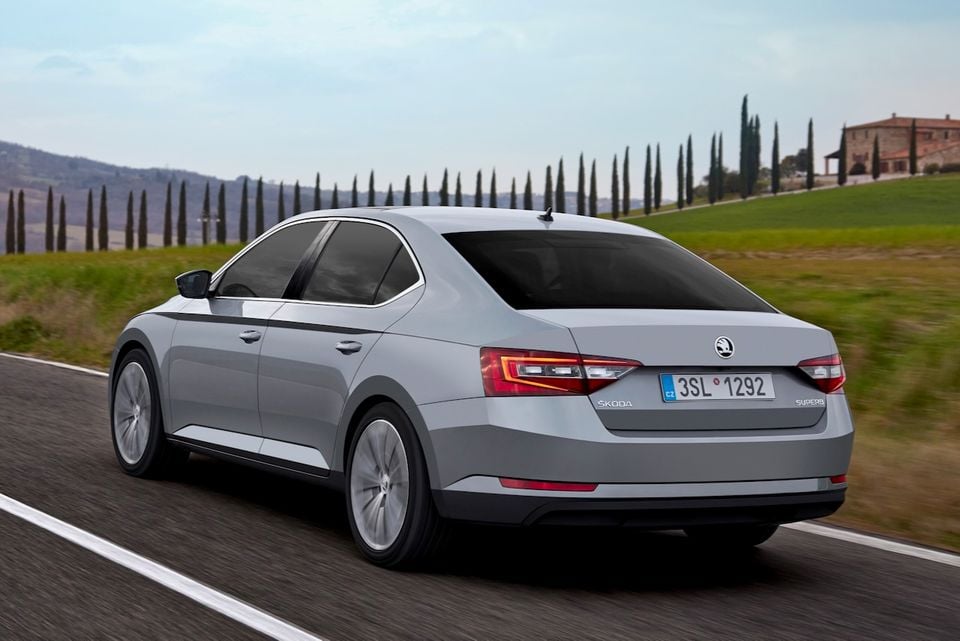 A silver Skoda Superb sedan driving on a road, seen from the rear three-quarter angle, with a scenic countryside backdrop.