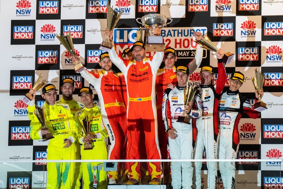 Racing drivers celebrate on the podium with trophies at the Bathurst 12 Hour, with Liqui Moly and NSW Government branding in the background.