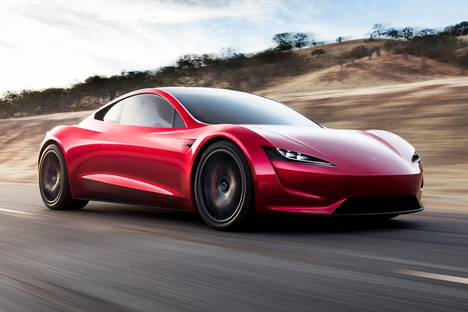 A red Tesla Roadster sports car driving along a road, seen from the front-side angle, with dry hills in the background