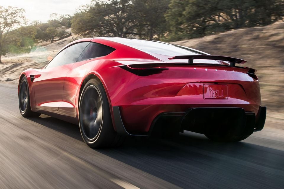 A red Tesla Roadster sports car seen from the rear three-quarter angle, driving on a road with trees in the background.