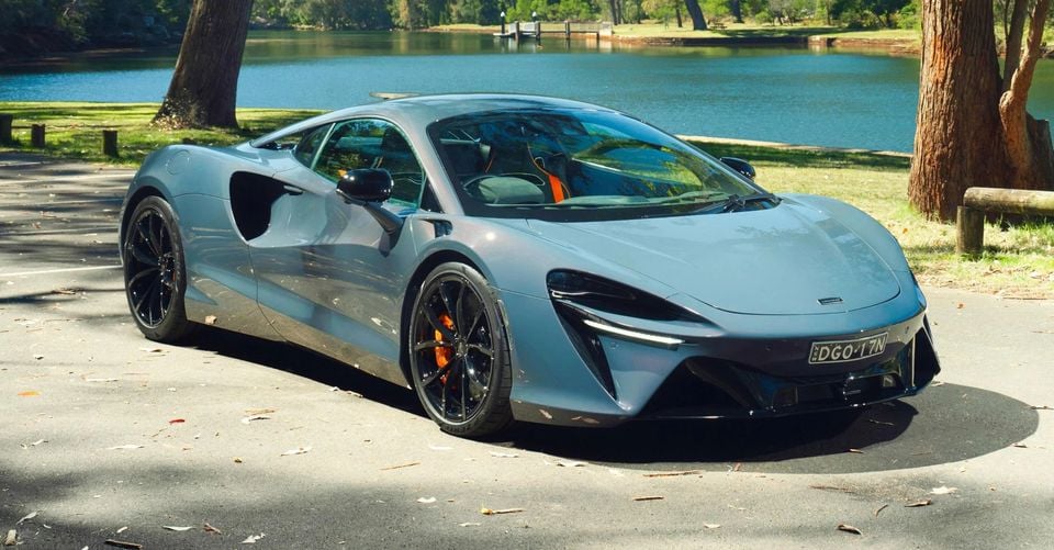 A grey McLaren Artura supercar seen from the front-side angle, parked near a lake in a park setting with an NSW number plate visible